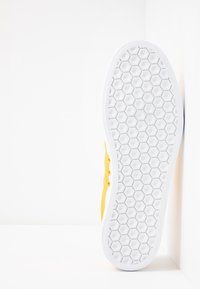 Gul sneaker med ett strukturerat tygöverdrag, vit gummisula med ett hexagonalt mönster och fyra öglor för snören.