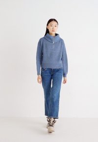 Lys blå ribbestrikket genser med høy krage og glidelås, kombinert med blå rette jeans og brune chunky sneakers. Enkelt og moderne design.