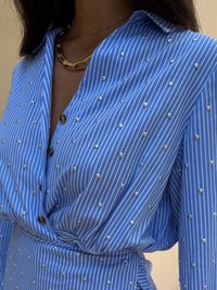 Camisa listrada azul e branca com tachas metálicas, apresentando um colarinho e fechamento com botão, feita de um tecido suave.