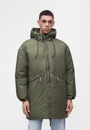 Young man wearing a long, olive green padded jacket with a hood and zipped pockets, standing against a plain light background.