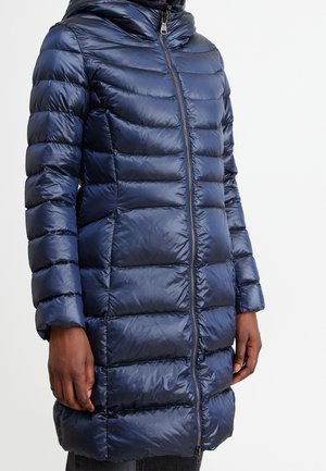 Henkilö, joka pukeutunut pitkässä, tummansinisessä tikatussa toppatakissa, jossa on etuvetoketju, seisoo yksinkertaisen vaalean taustan edessä.