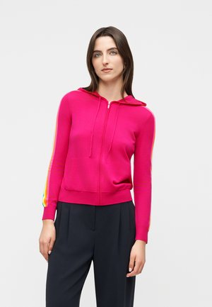 Frau trägt einen leuchtend pinken Reißverschluss-Hoodie mit einem Regenbogenstreifen an den Ärmeln und schwarze plissierte Hosen, steht vor einem weißen Hintergrund.