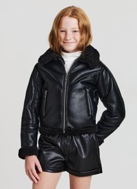 Schwarze Kunstleder-Bomberjacke mit einem flauschigen Kragen und Bündchen, mit Reißverschluss versehenen Fronttaschen und elastischem Saum. Getragen mit glänzenden schwarzen Shorts.