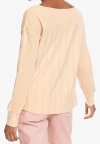 Chemise beige clair à manches longues avec un design côtelé texturé, épaules tombantes et un large décolleté, associée à un pantalon rose clair.