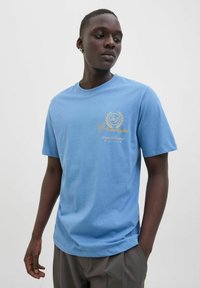 Tricou din bumbac de culoare albastru deschis, cu guler rotund, mâneci scurte și un imprimeu cu logo auriu pe piept. Textură netedă și croială lejeră.