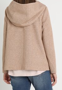 Beige uldblandingsjakke med hætte, der har en afslappet pasform, synlige sømme og en tekstureret overflade. Båret over en lys topp og jeans.