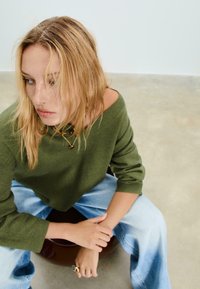 Blonde vrouw zittend op een kruk, gekleed in een groene trui, lichtblauwe jeans, gelaagde gouden kettingen en een grote ring, kijkend opzij.