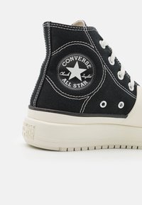 Baskets montantes noires Converse Chuck Taylor All Star avec coutures blanches, semelle en caoutchouc blanche et patch de marque à la cheville.