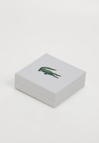 Bijela kutija kvadratnog oblika s teksturiranom površinom, s zelenim logotipom krokodila na vrhu. Kutija ima elegantan, minimalistički dizajn.