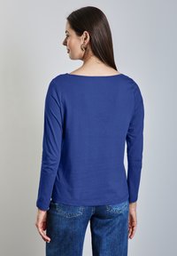T-shirt à manches longues en tissu bleu vif, avec un décolleté large et une texture lisse. Associé à un jean en denim, offrant une coupe décontractée.