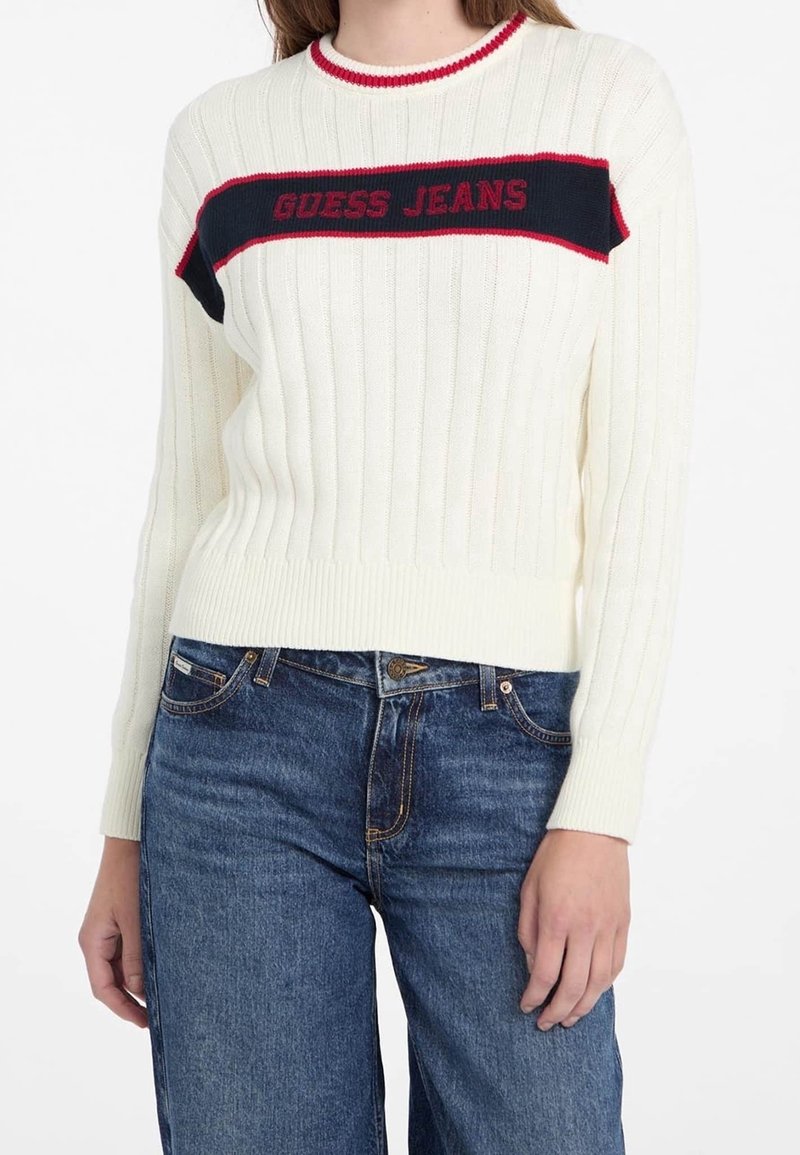 Femme portant un pull blanc côtelé avec une bande "GUESS JEANS" bleu marine et rouge sur la poitrine et un jean taille haute bleu.