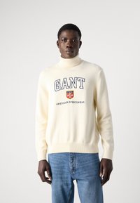 Cremfarbener Rollkragenpullover mit blauer Aufschrift "GANT USA AMERICAN SPORTSWEAR" auf der Vorderseite, kombiniert mit hellblauen Jeans.