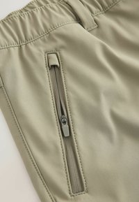 Bikses gaiši bēšā materiālā ar gludu tekstūru, ar leņķveida rāvējslēdzēja kabatu augšstilbā drošai uzglabāšanai.