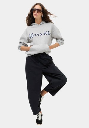 Kvinde iført store solbriller, grå hættetrøje med teksten "Marseille", sorte vidde bukser, sort-hvide sneakers, stående med det ene ben løftet.