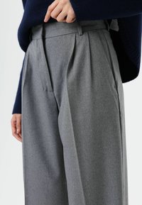 Gray pleated trousers with a high waist, made of soft material. Features side pockets and a smooth texture. Worn with a navy sweater.