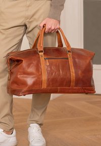 Brun læder rejsetaske med brune læderdetaljer, dobbelte håndtag, lynlåsåbning i toppen og sidelomme; har en glat overflade og en robust konstruktion.