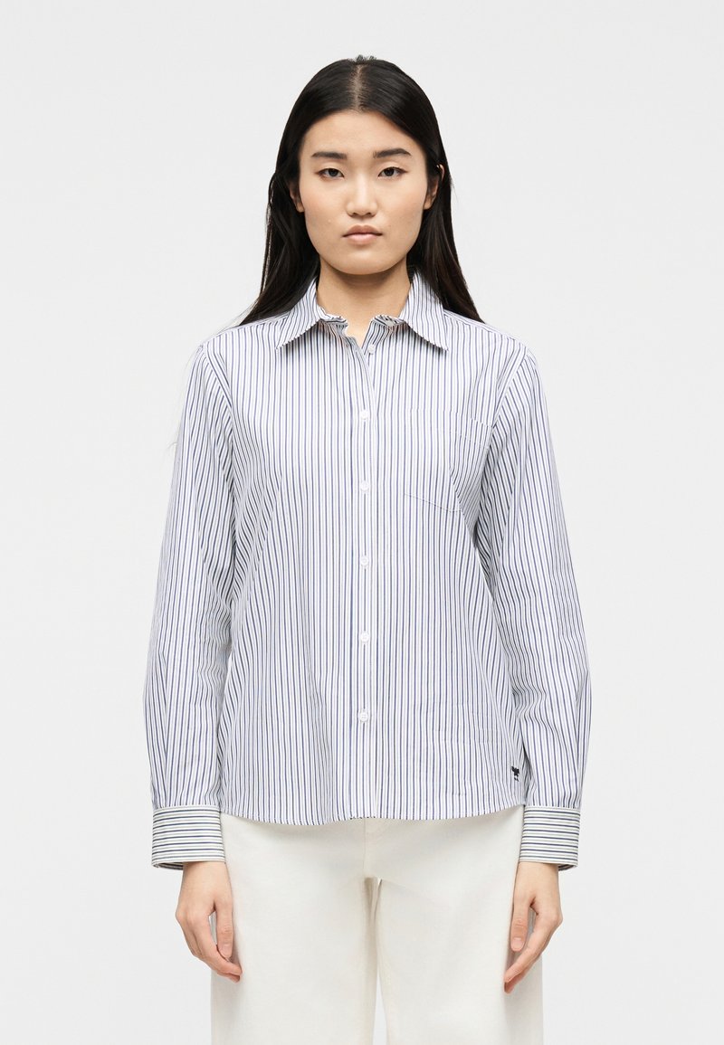 Γυναίκα που φοράει λευκό πουκάμισο με λεπτές μπλε κάθετες ρίγες και αντίθετες ριγωτές μανσέτες, συνδυασμένο με λευκό παντελόνι, στέκεται όρθια.