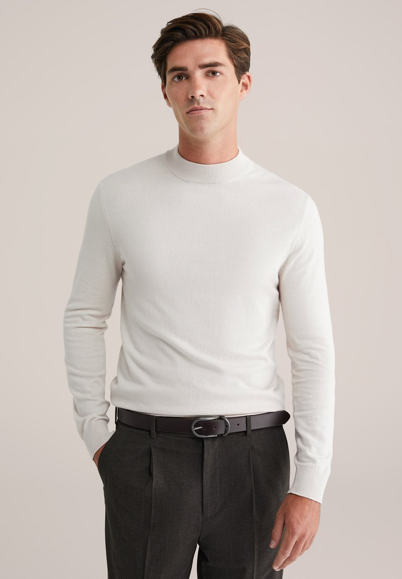 Valge pika varrukaga sviiter kõrge kaelusega, kombineeritud tumedate hallide pükste ja musta naha rihmaga. Pehme tekstuur, keha lähedane disain.