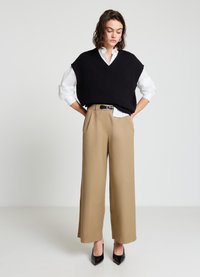 Schwarze Strickweste mit V-Ausschnitt über einem weißen Hemd; beige Weitbein-Hose; schwarze spitze Schuhe; minimalistisches Design.