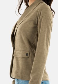 Blazer caqui con una tela texturizada, que presenta un cuello chal, un detalle de un solo botón y un bolsillo lateral. Ajuste entallado con mangas largas.