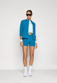 Ensemble de veste et short bleu vif, avec un top blanc court, en matériaux élastiques, texture lisse, et accents du logo Nike.