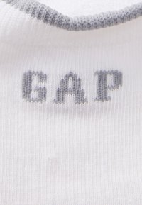 Hvit sokk med grå ribbestrikket mansjett, med ordet "GAP" i grå strikkede bokstaver på siden, laget av teksturert bomullsblanding.