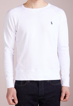 Homme portant un t-shirt blanc à manches longues avec un petit logo sombre sur la poitrine et un jean bleu foncé, debout les bras détendus sur les côtés.