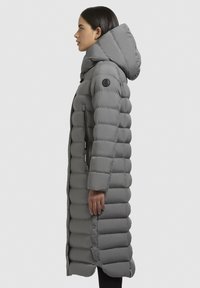 Lang, fyldig grå parka med hætte, der har vandret baffle, en lynlåslomme på siden og et logo-patch på venstre ærme.