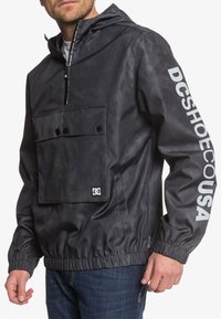 Black pullover jacket with a hood, featuring a front pocket, elastic cuffs, and DC logo printed in white on the sleeve. Smooth texture.