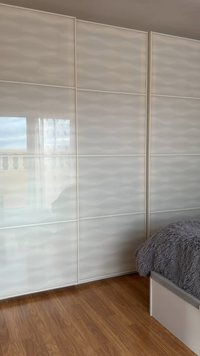 Λευκή ντουλάπα με πάνελ και γυαλιστερό φινίρισμα που αντανακλά το παράθυρο και το μπαλκόνι, δίπλα σε ένα κρεβάτι με γκρι χνουδωτή κουβέρτα πάνω σε ξύλινο πάτωμα.