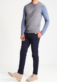 Pull sans manches gris porté sur une chemise à manches longues bleue, pantalon bleu marine et chaussures beige clair. Présente un col en V et des textures simples.