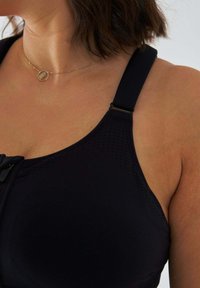 Czarny sportowy biustonosz z szerokimi paskami, teksturowaną tkaniną i przednim zamkiem błyskawicznym. Zawiera niewielki metalowy element ozdobny przy pasku.