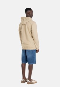 Beige Kapuzenpullover mit Kapuze, kombiniert mit blauen Jeansshorts. Die Person trägt braune und weiße Sneakers. Sichtbare Nähte und Taschen an den Shorts.