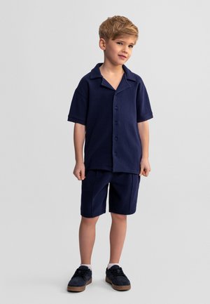Jeune garçon aux cheveux clairs portant une chemise à manches courtes bleu marine avec boutons et un short assorti, debout devant un fond clair uni.