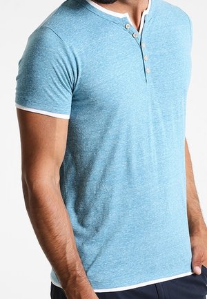 Mężczyzna w jasnoniebieskiej, krótkorękawowej koszulce henley z białym wykończeniem i trzema beżowymi guzikami, stojący na tle jednolitej białej ściany.