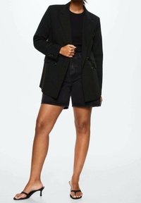 Blazer noir oversize avec fermeture croisée à boutons, porté sur un haut noir et un short en denim taille haute, associé à des sandales slides noires.