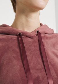 Felpa morbida in fleece color malva con collo a coulisse, caratterizzata da nastri bordeaux in satin e occhielli, con cuciture dettagliate vicino al cappuccio.