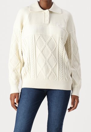 Femme portant un pull en maille crème avec des motifs torsadés et losanges, un col avec trois boutons, associé à un jean bleu foncé.