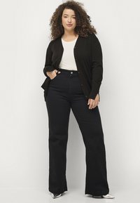 Zwarte cardigan met knopen over een witte shirt, gecombineerd met hoge taille, flared, donkere jeans en zilveren puntige schoenen.
