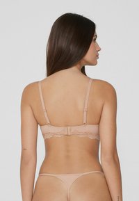 Femme aux longs cheveux noirs portant un soutien-gorge en dentelle nude et un string assorti, vue de dos sur un fond clair uni.