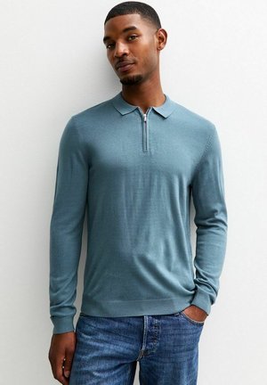 Suéter de polo de manga larga azul claro con cuello con cremallera y puños acanalados, combinado con jeans de mezclilla. Textura suave y diseño ajustado.