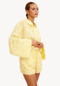 Mujer con una camisa de manga larga texturizada en amarillo pálido y pantalones cortos a juego con cordón, de pie con las manos entrelazadas, mirando hacia la derecha.