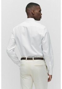Chemise blanche à manches longues avec une coupe ajustée, associée à un pantalon de couleur claire. La chemise présente un empiècement de dos structuré et des poignets boutonnés.