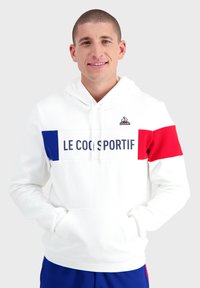 Sweat à capuche blanc avec des rayures horizontales bleues et rouges, arborant "LE COQ SPORTIF" en lettres majuscules, poches kangourou et capuche ajustable avec cordon.