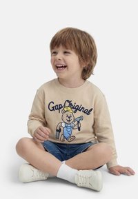 Χαμογελαστός νεαρός αγόρι με καστανά μαλλιά κάθεται σταυροπόδι, φοράει μπεζ φούτερ με γραφικό αρκούδα, τζιν σορτς, άσπρες κάλτσες και λευκά αθλητικά παπούτσια.