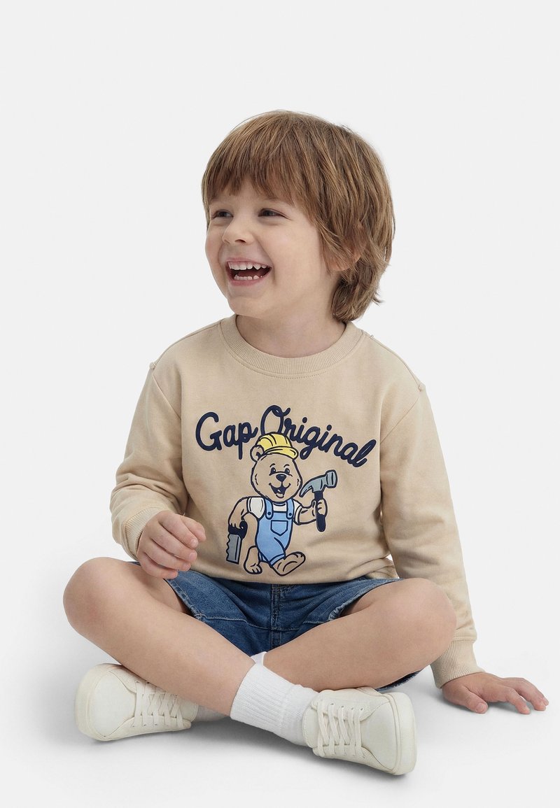 Χαμογελαστός νεαρός αγόρι με καστανά μαλλιά κάθεται σταυροπόδι, φοράει μπεζ φούτερ με γραφικό αρκούδα, τζιν σορτς, άσπρες κάλτσες και λευκά αθλητικά παπούτσια.