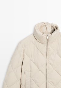 Gevoerde beige jas met een hoge kraag, ritssluiting aan de voorkant en zijzakken. Gladde textuur en diagonale stiksels voor isolatie.