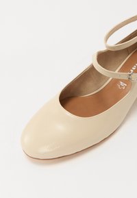 Beige lackläderballettsko med rund tå, ankelsnöra och brun vadderad innersula. Slät yta med minimala designdetaljer.