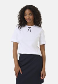 Hvid kortærmet t-shirt med rund halsudskæring og sort sløjfeaccent på brystet, kombineret med en marineblå nederdel. Glat tekstur, afslappet pasform.