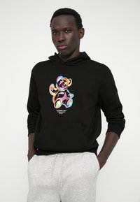 Felpa nera con grafica di un orso in colori vivaci; presenta una tasca frontale e cappuccio con stringhe, abbinata a pantaloni della tuta grigi.